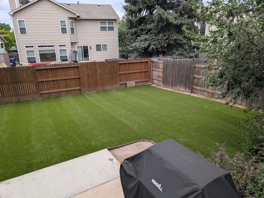 Lush green artificial turf lawn in a backyard with wooden fence and nearby barbecue grill.