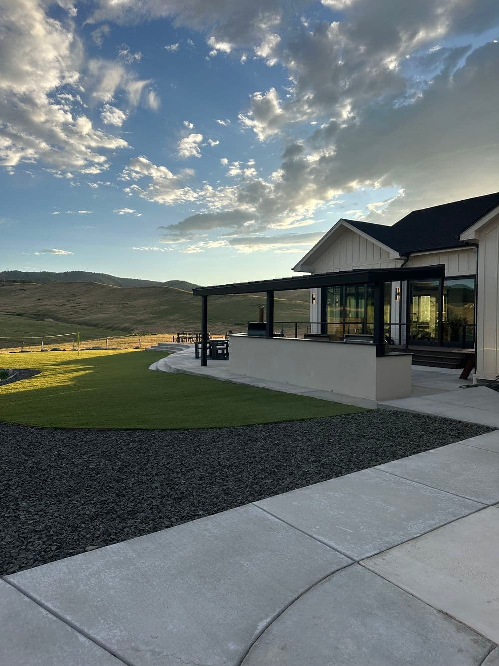 Modern house exterior with patio, green lawn, and scenic countryside view under a cloudy sky.