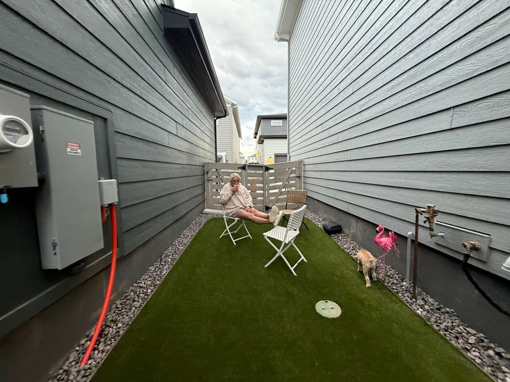 Person sitting on a chair in a narrow yard between two gray houses with a dog nearby.