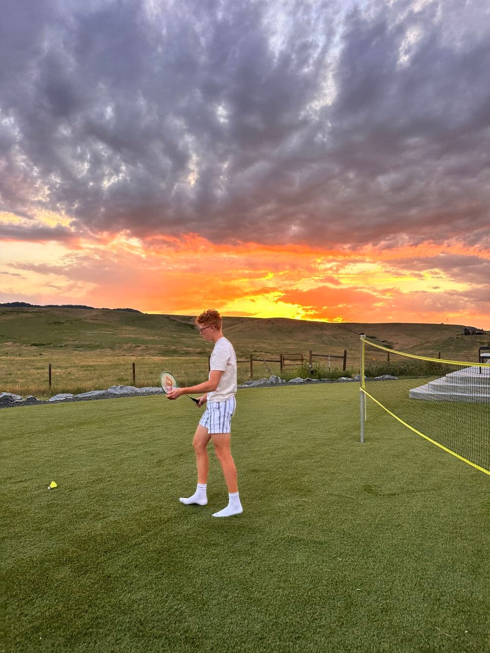 Boy playing tennis on a grassy court at sunset, with vibrant clouds in the background.