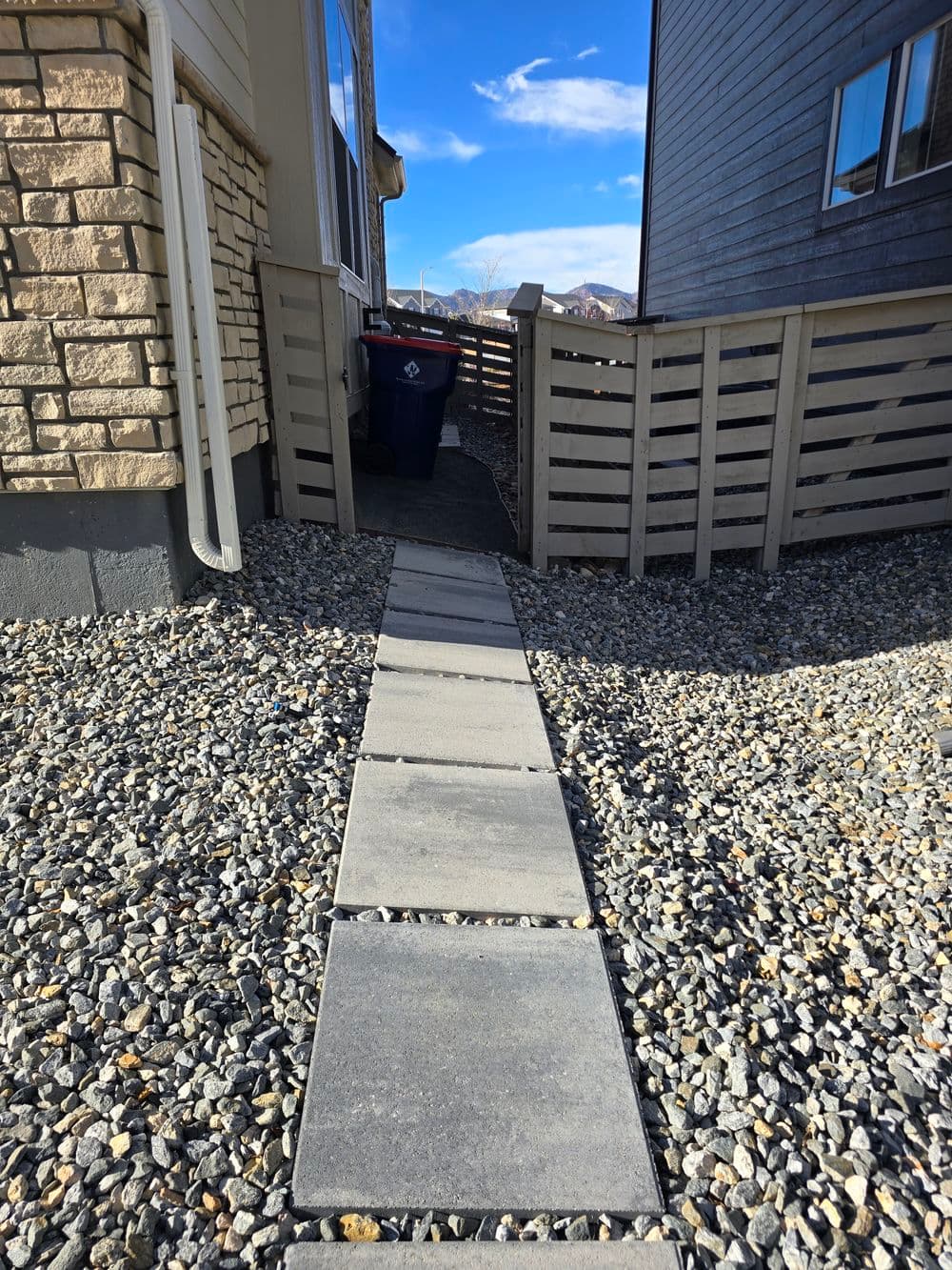 Pathway of gray stone tiles leading to a fenced area with a trash bin and blue sky above.