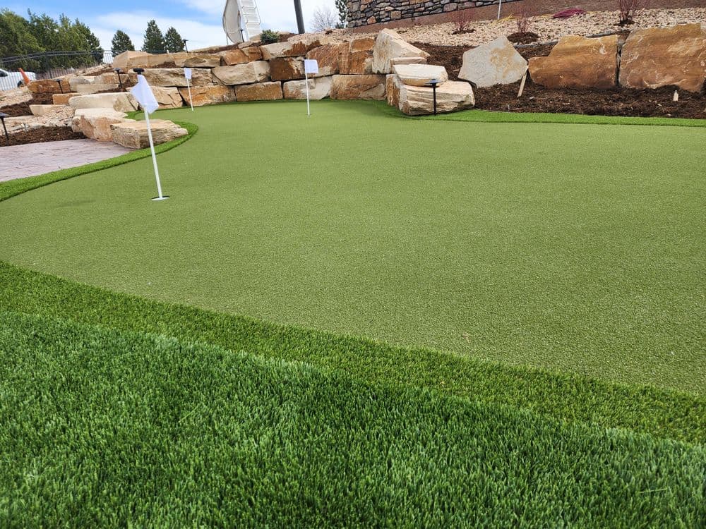 Artificial turf putting green with golf flags and rock landscaping in a landscaped area.
