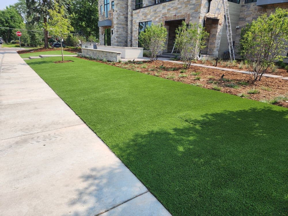 Lush green artificial turf alongside freshly landscaped garden and stone building entrance.